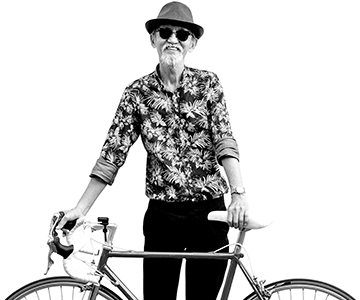 A person wearing a fedora and sunglasses smiles and stands behind a bicycle