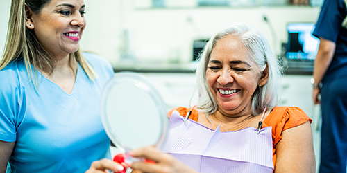 One individual in a dentist's office holds up a mirror while the other stands besides them